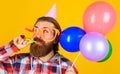 Bearded man in party hat and glasses with balloons blowing noisemaker. Birthday holiday celebration.