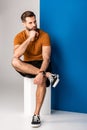 Bearded man in shorts and brown polo sitting on white cube on grey and blue