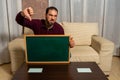 Bearded man sitting on the couch at home with a blackboard with his thumb down