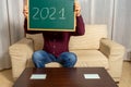 Bearded man sitting on the couch at home with a blackboard with 2021 written on it as a sing of approval of an exciting year