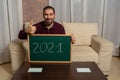 Bearded man sitting on the couch at home with a blackboard with 2021 written on it and his thumbs up in approval of an exciting