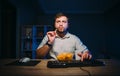 Bearded man sitting at night at the computer for dinner chips, with a serious face looking at the camera