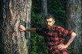 Tall brutal man standing in forest. Lumberjack resting while leaning on tree. Hipster with roses tattoo on arm Royalty Free Stock Photo