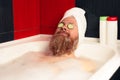 Bearded man taking bath with head wrapped in towel and cucumber slices on his eyes. Funny hipster relaxing in bathtub Royalty Free Stock Photo