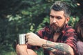 Bearded man with tea or coffee cup in forest. Tourist in plaid shirt hold mug. Hipster with long beard relax on natural