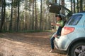 Bearded man tourist in felt hat with hot tea in thermos sitting in car trunk in pine forest. Road trip, weekend on nature, outdoor