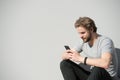 Bearded man use mobile phone on grey wall. Macho with smartphone on sunny outdoor. Guy with beard and blond hair in casual tshirt