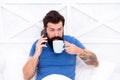 Bearded man using mobile technology in bed. Handsome guy talking on phone and drinking coffee at home. Modern life new Royalty Free Stock Photo