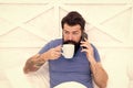 Bearded man using mobile technology in bed. Handsome guy talking on phone and drinking coffee at home. Modern life new Royalty Free Stock Photo