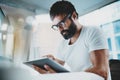 Bearded man in white tshirt wearing eye glasses and using portable electronic pro tablet computer at modern lightful