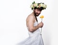 Bearded man in a woman`s wedding dress on her naked body, holding a flower. on his head a wreath of flowers. funny