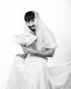 Bearded man in a woman`s wedding dress on her naked body, holding a flower. on his head a wreath of flowers. funny Royalty Free Stock Photo