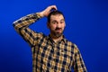 Bearded man in a yellow plaid shirt scratching his head with doubt gesture isolated on blue studio background