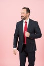 Bearded manager holds hand on flap of blue suit jacket wearing red tie on white shirt isolated over pink background
