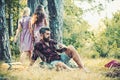 Bearded man drinking coffee or tea by campfire. Two girls in retro dresses walking on path in woods, turn back