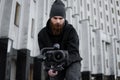 Bearded Professional videographer in black hoodie holding professional camera on 3-axis gimbal stabilizer. Filmmaker