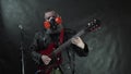 Bearded rockstar in transparent mask, respirator and leather jacket plays red electric guitar on stage with black