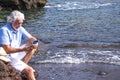 Bearded senior man with white hair using smart phone sitting on the rocks near the water enjoying the vacation at sea - concept of Royalty Free Stock Photo