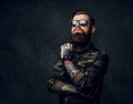 Bearded tattooed guy in military shirt and sunglasses posing with thoughtful look. Studio photo against dark wall Royalty Free Stock Photo