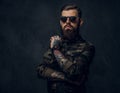 Bearded tattooed guy in military shirt and sunglasses posing with thoughtful look. Studio photo against dark wall