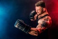 Bearded tattooed sportsman muay thai boxer in black undershirt and boxing gloves fighting on dark background with smoke.