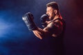 Bearded tattooed sportsman muay thai boxer in black undershirt and boxing gloves fighting on dark background with smoke.