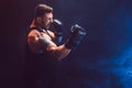 Bearded tattooed sportsman muay thai boxer in black undershirt and boxing gloves fighting on dark background with smoke.