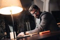 Bearded young businessman in glasses sits by the wooden table with lamp and using contemporary mobile notebook in loft office at n