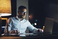 Bearded young businessman wearing white shirt working on modern loft office at night. Man using contemporary notebook Royalty Free Stock Photo