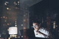 Bearded young businessman working on modern loft office at night. Man using contemporary notebook texting message, holding cup esp