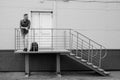 Bearded young man in jeans and denim shirt is standing on porch of industrial building near closed door. job search or firing