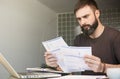 Bearded young man sitting at table looking at documents and thinking. Business man going through paperwork at home