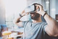 Bearded young man wearing virtual reality goggles in modern coworking studio. Smartphone using with VR headset in office