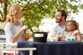 Beardy caucasian male with little girl in his lap looking at blonde female, smiling, sitting outdoor