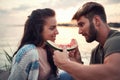 Beardy man feeding a woman with a slice of a watermelon by the water