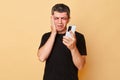 Beaten traumatized man in black T-shirt with bruises and abrasions on his face isolated over beige background holding smartphone