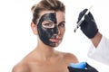 Beautician`s hands in medical gloves hold a cosmetic brush and apply a black charcoal purifying mask to the face of an attractive