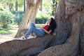 A beautiful, adult, dark-haired, curly-haired woman with the features of a Latin gypsy is lying on the roots of a giant ficus tree