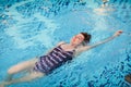 Beautiful adult woman in striped swimsuit swimming in blue pool on her back at resort. Sport activity health concept.