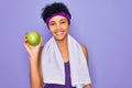 Beautiful african american afro sporty woman doing exercise wearing towel eating green apple with a happy face standing and Royalty Free Stock Photo