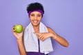 Beautiful african american afro sporty woman doing exercise wearing towel eating green apple very happy pointing with hand and Royalty Free Stock Photo