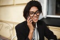 Beautiful African American girl in glasses sitting at the table of cafe and talking on her cellphone. Pretty lady with