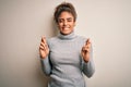 Beautiful african american girl wearing turtleneck sweater standing over white background gesturing finger crossed smiling with Royalty Free Stock Photo