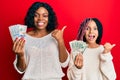 Beautiful african american mother and daughter holding dollars pointing thumb up to the side smiling happy with open mouth Royalty Free Stock Photo