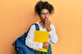 Beautiful african american woman with afro hair wearing school bag and holding books covering mouth with hand, shocked and afraid Royalty Free Stock Photo