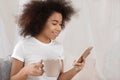 Beautiful African-American woman drinking tea while using mobile phone at home Royalty Free Stock Photo