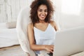 Beautiful african girl in sleepwear smiling looking at camera sitting in chair with laptop at home. Copy space. Royalty Free Stock Photo