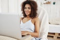 Beautiful african girl in sleepwear smiling looking at camera sitting in chair with laptop at home. Copy space. Royalty Free Stock Photo