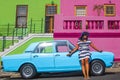 A beautiful African woman in a blue and white striped dress in front of a vintage Ford Cortina and traditional homes of Bo-Kaap, C