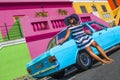 A beautiful African woman in a blue and white striped dress in front of a vintage Ford Cortina and traditional homes of Bo-Kaap, C